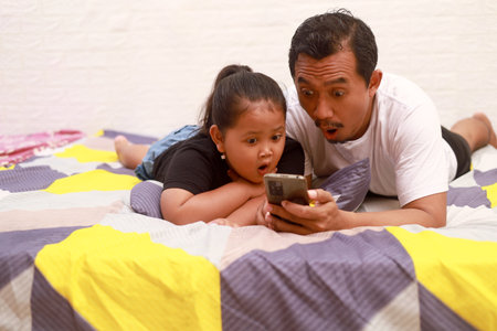 Surprised Asian dad and his daughter lie on the bed at home while watching something on cell phoneの写真素材