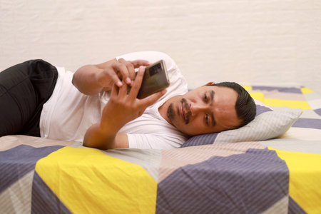 Asian man lie on the bed at home while holding cell phoneの写真素材