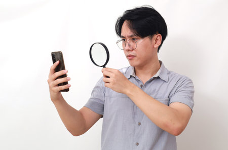 Curious asian man looking his cell phone through magnifying glass. Isolated on whiteの写真素材