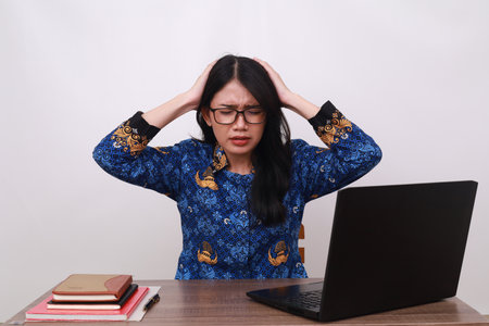 Stressed Asian female in batik korpri, indonesian traditional uniform holding her head while workingの写真素材