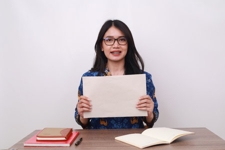 Happy Asian female in batik korpri, indonesian traditional worker uniform showing a blank paperの写真素材
