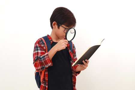 Happy smart asian student holding a book while looking through a magnifying glass. Isolated on whiteの写真素材