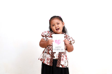 Adorable asian little girl in batik showing happy fathers day greeting card. Isolated on white...