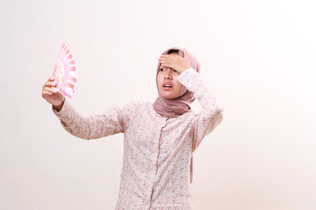 Person in modest dress standing while holding Indonesian banknotes, showing stressed expression. Isolated on whiteの写真素材