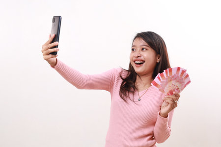 Wow excited young asian girl getting prize from her phone while holding Indonesia banknotes. Isolated on whiteの写真素材