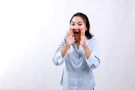Carefree asian enthusiastic woman shouting long distance, calling friend, breaking free emotions, yelling out loud, hold hands near opened mouth, searching someone in crowd, white backgroundの写真素材