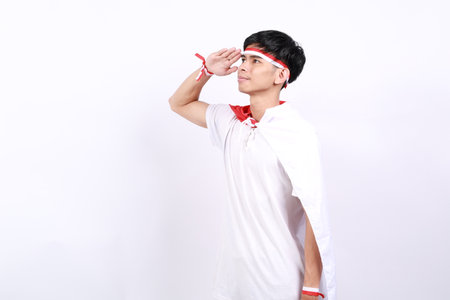 Asian young boy with red white ribbon for celebrating Indonesia independence day standing with salute hand gesture while looking side aboveの写真素材