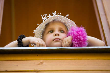 The little girl in a summer hat looks out of a window of a wooden country houseの写真素材