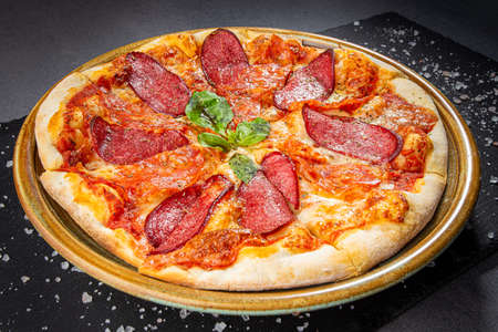 Italian salami pizza with cheese sauce and crispy pepper with roasted spice mix, wooden dish plate on a stone table top.の写真素材