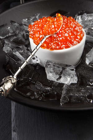 Red caviar in a white ceramic bowl with a spoon and some ice in a dish. Dark background, top viewの写真素材