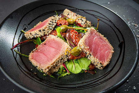 Close up of tuna slices withsesam seeds and fresh vegetables on a dark plate.の写真素材