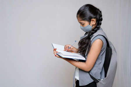 The Indian lovely adorable charming little girl is looking down at the copybook in her hands and writing information there, she is wearing a dress, isolated on bright plane background, copy spaceの写真素材
