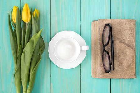 Old book with glasses,yellow tulips and empty cup on wooden backgroundの写真素材