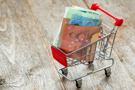 Consuming concept with shopping cart and euro currency on wooden backgroundの写真素材