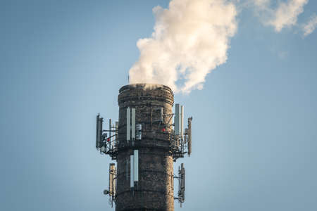 Top of high industrial chimney with multiple cell phone antennas, smoke comming out on clear blue...
