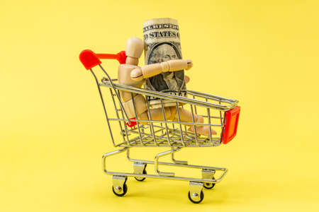 Wooden man in a shopping cart with one dollar banknote. Shopping concept.