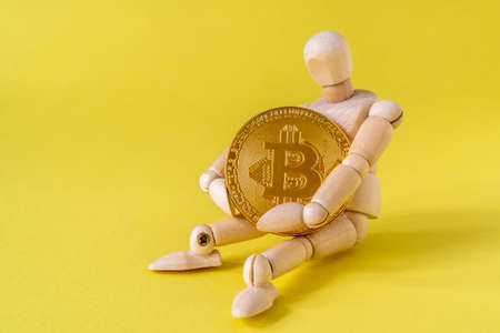 Wooden doll tightly holding Bitcoin while sitting on yellow backgroundの写真素材
