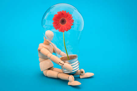 Wooden man holds a  light bulb with red flower inside. Concept of green energy