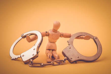 Wooden man with handcuffs on hands sitting on yellow background. Conceptual image.の写真素材