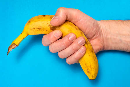 Male hand holding a banana on a blue background. Minimal fruit concept.の写真素材