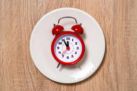 Plate with alarm clock with five minutes to twelve o'clock on the dial. Lunch time conceptの写真素材