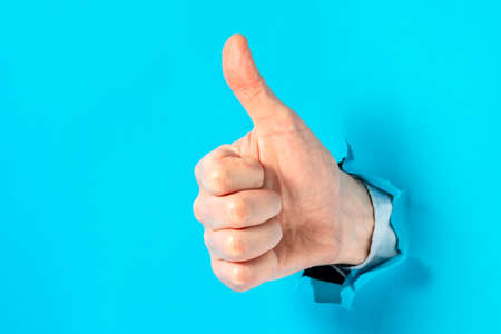 Close up of man hand with thumb up gesture through blue hole in paper wall. Copy space.の写真素材