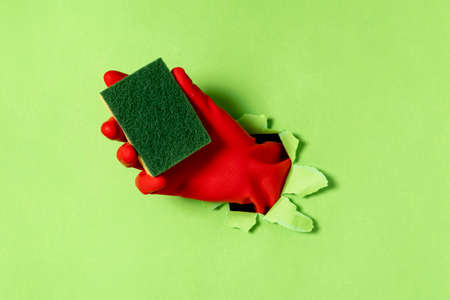 A hand in a rubber glove with dishwashing sponge sticks out of a torn hole in green paper. Cleaning concept.の写真素材