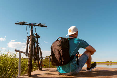Back view of unrecognizable man with backpack sitting at bicycle on wooden pier on sunny day. Active holiday concept.の写真素材