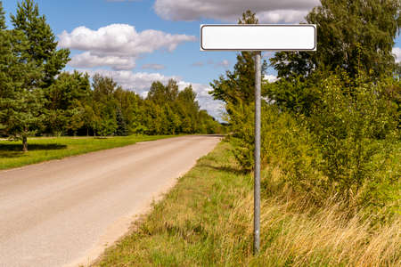 Road with white empty signpost on roadside at city entrance against sunny landscape background.