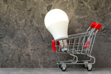Shopping cart with bright glowing light bulb on grey granite background. Energy saving, energy prices concept.の写真素材