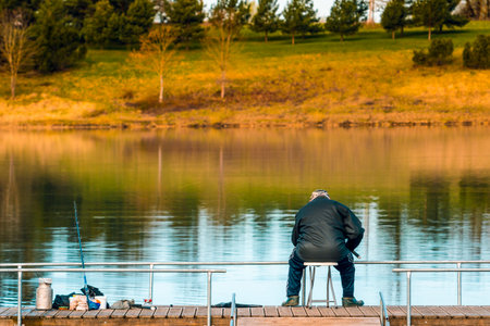 Senior man sitting at the lake pier with rods and fishing equipment in the morning. Back view of senior man fishing in a lake.の写真素材