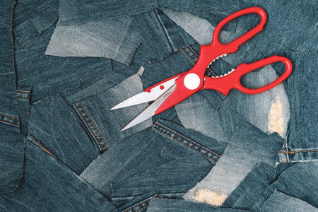 Red scissors lie on denim jeans patching pieces. Denim jeans background repair. Recycle jeans fashion conceptの写真素材