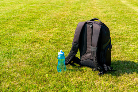 Black backpack and water bottleon the green grass background. Traveling, hiking concept.
