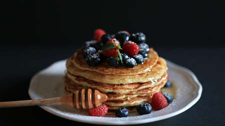 Stack of pancakes with honey, blueberries and red raspberries with icing sugar on a dark background