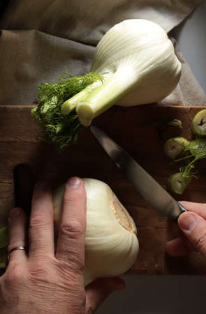 Close-up of a man's hand slicing fennel on a wooden cutting board. Dark background. Directly above.の写真素材
