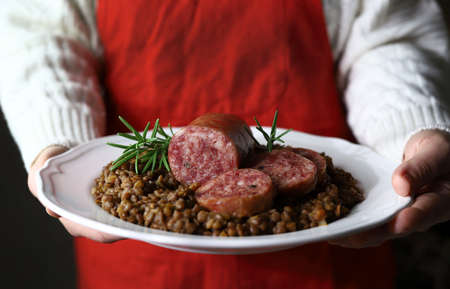 Italian food. Hands holding white plate with Italian large pork sausage cotechino served with lentils. Close-up.の写真素材