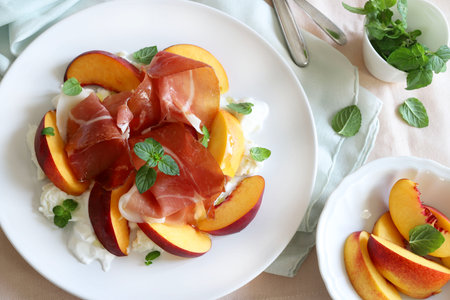 A delicious salad with peach, ham and burrata cheese, seasoned with olive oil and mint leaves in a white plate on kitchen table background.の写真素材