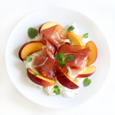 A delicious salad with peach, ham and burrata cheese, seasoned with olive oil and mint leaves in a white plate on kitchen table background.の写真素材