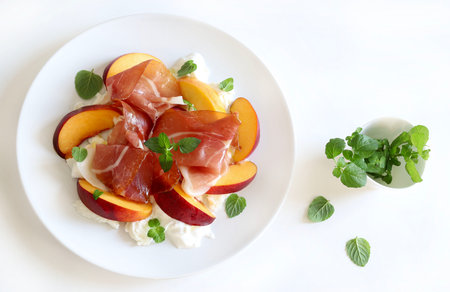 A delicious salad with peach, ham and burrata cheese, seasoned with olive oil and mint leaves in a white plate on kitchen table background.の写真素材