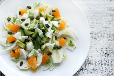 Fresh salad of fennel, oranges, avocado, pieces of cheese and pumpkin seeds dressed with extra virgin olive oil on a white wooden background.の写真素材