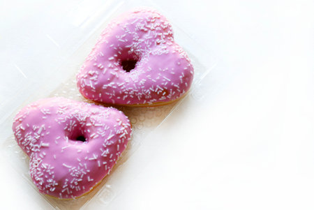 Two heart-shaped donuts with pink icing and sprinkles, in a clear plastic container on a white background. Romantic or Valentine's Day theme.の写真素材