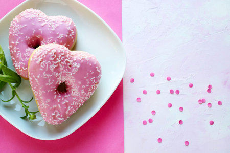 Two heart-shaped donuts with pink icing and sprinkles on a white plate against a bright pink background. Romantic or Valentine's Day theme.の写真素材