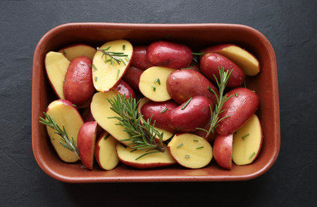 Raw red potatoes, cut in half, with rosemary sprigs and olive oil, ready to be roasted on a black background. Healthy and vegetarian food. Overhead view.の写真素材