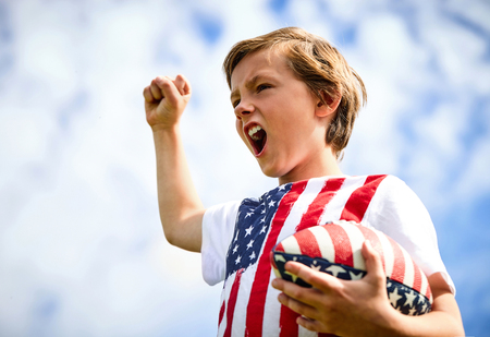 Portrait of ecstatic little rugby player of junior team wearing uniform with stars and stripes holding ball and screaming with winning gestureの写真素材