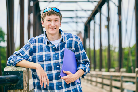 Portrait of cheerful young Caucasian male student wearing checked shirt and sunglasses standing on bridge, holding folder, looking at camera and smiling