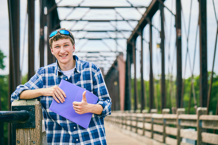 Portrait of positive young Caucasian male student wearing checked shirt and sunglasses standing on bridge, holding folder, showing thumbs up and smilingの写真素材