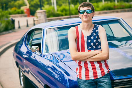 Portrait of successful teenage Caucasian boy wearing American tank top and sunglasses standing at his new blue car with crossed arms and smilingの写真素材