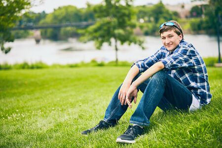 Portrait of smiling young Caucasian man wearing checked shirt and sunglasses sitting on grass,...