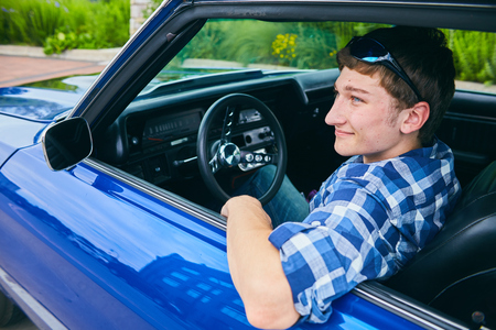 Portrait of positive teenage boy wearing checked shirt sitting in car, looking away and smilingの写真素材