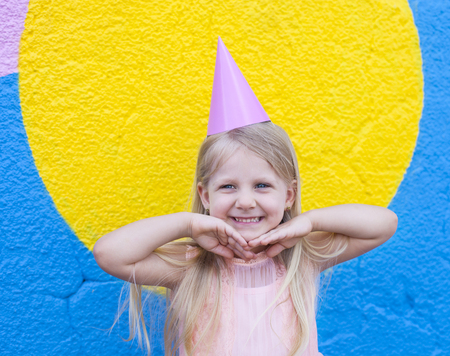 Happy little girl with birthday cap on head holding hands next to her chin.の写真素材